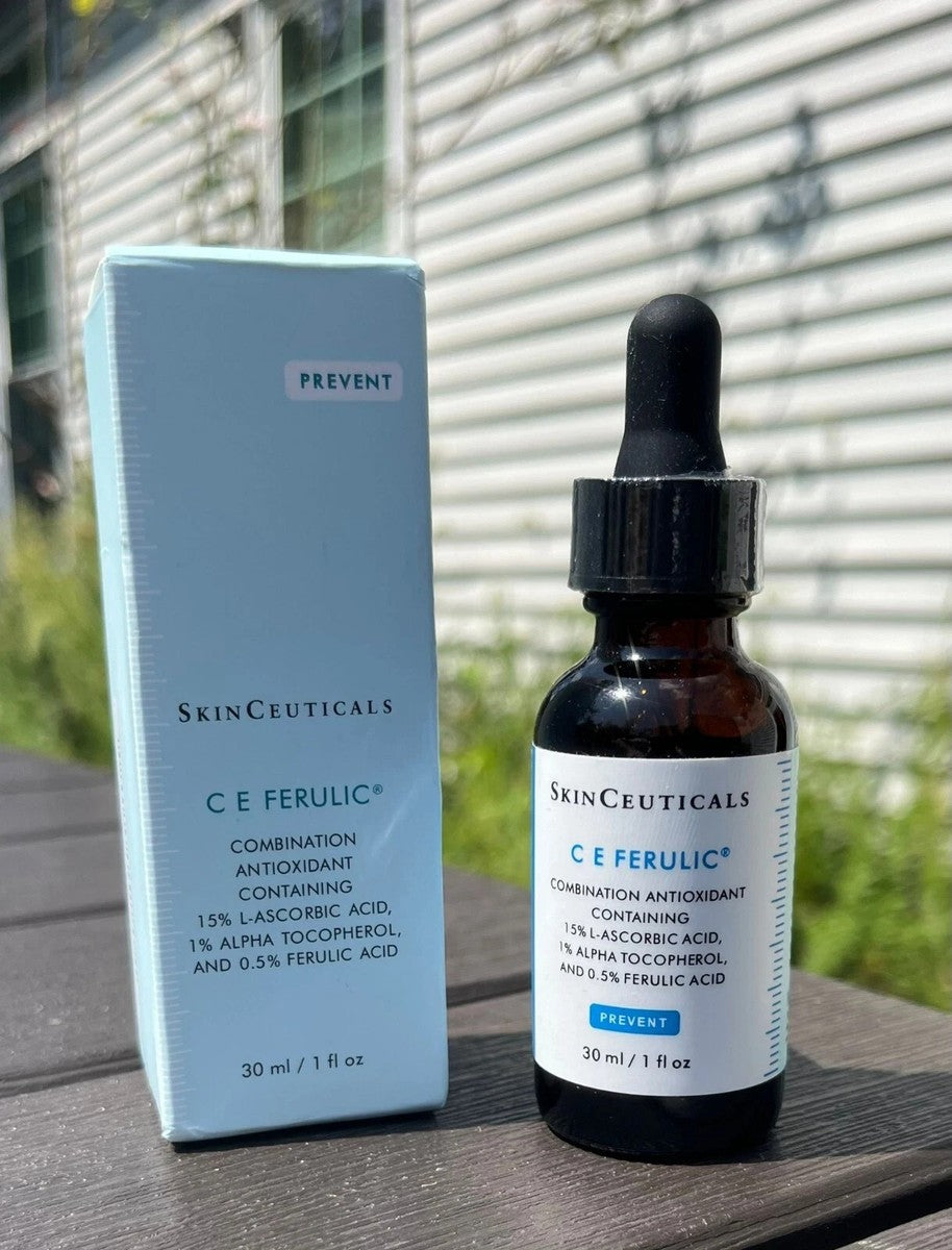 SkinCeuticals C E Ferulic combination antioxidant serum in an amber glass dropper bottle next to its light blue packaging box, displayed on a dark wooden surface with a blurred outdoor patio background.